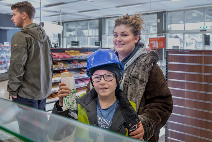 En pojke i blå arbetshjälm håller i en varmkorv, i bakgrunden hans mamma och ytterligare en manlig kund