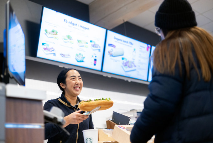 En kvinnlig butiksanställd håller fram en smörgås med serveringstång till en kund i förgrunden