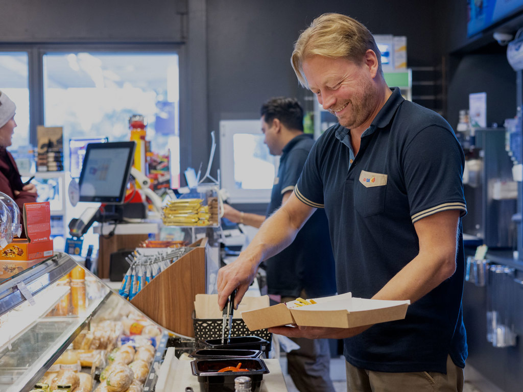 En man i arbetskläder förbereder mat bakom disken på en OKQ8 station, 